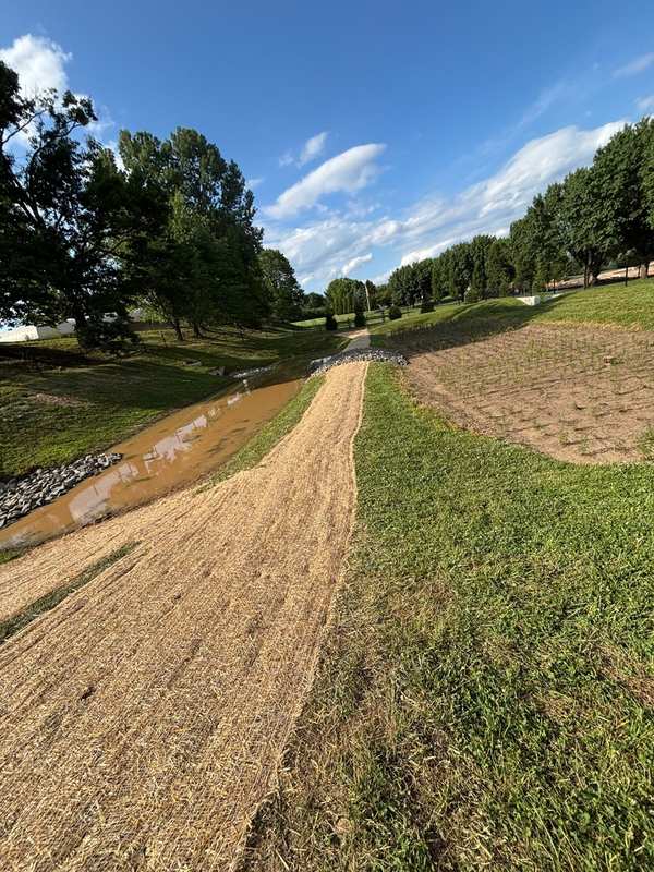 A pond overflow under repair: new riprap, an erosion-mat chute down the rebuilt berm and a freshly seeded slope